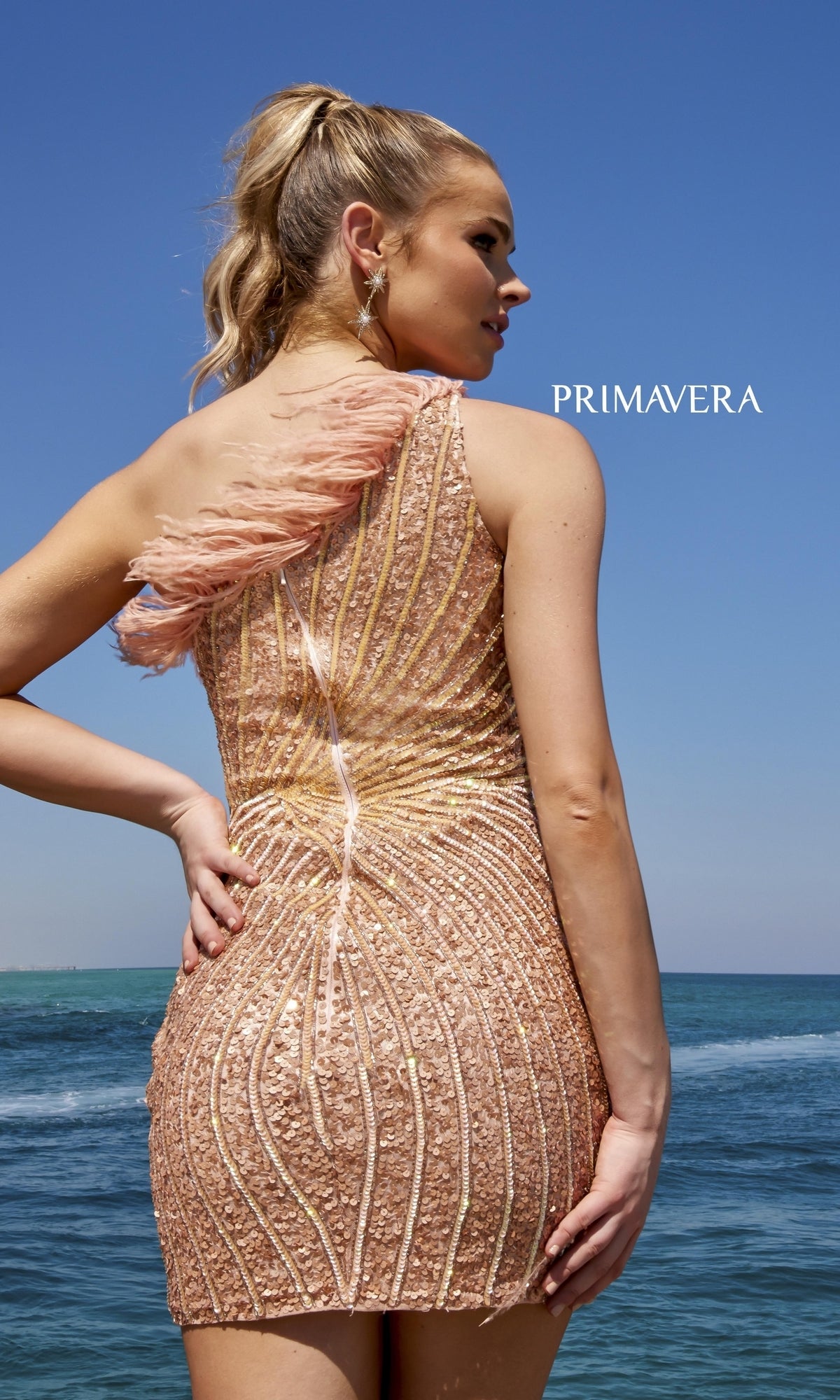 A woman in the One-Shoulder Sequin Party Dress with Feathers 4002 stands by the ocean under a clear blue sky. The shimmering gold mini dress sparkles in the sunlight, with